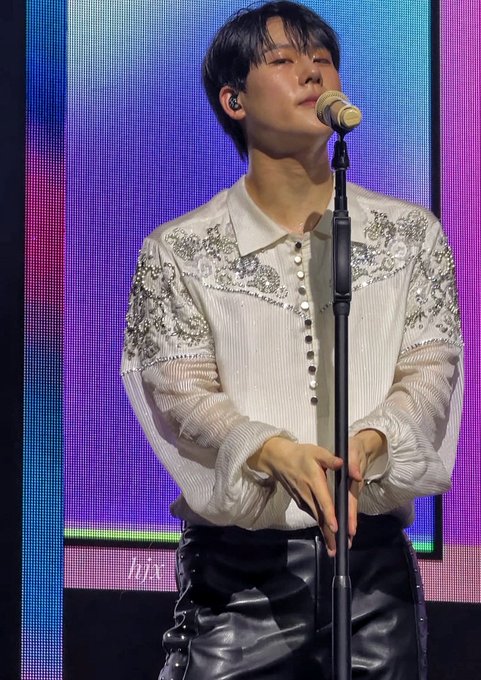 A male performer with short dark hair wearing an embellished white shirt and black leather pants stands at a microphone on stage during a concert.