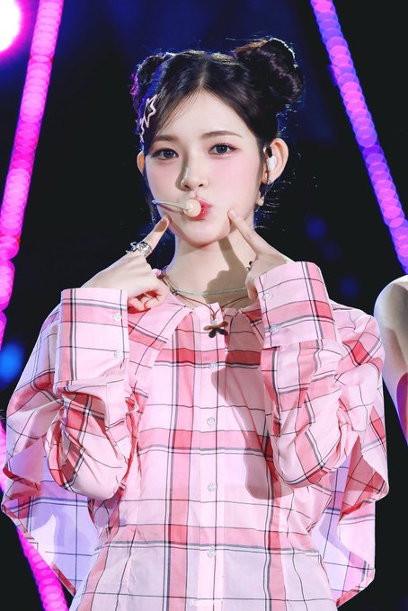 A young woman with dark hair styled in space buns, wearing a pink plaid blouse and a headset microphone, poses on a stage with vibrant pink and blue neon lighting.