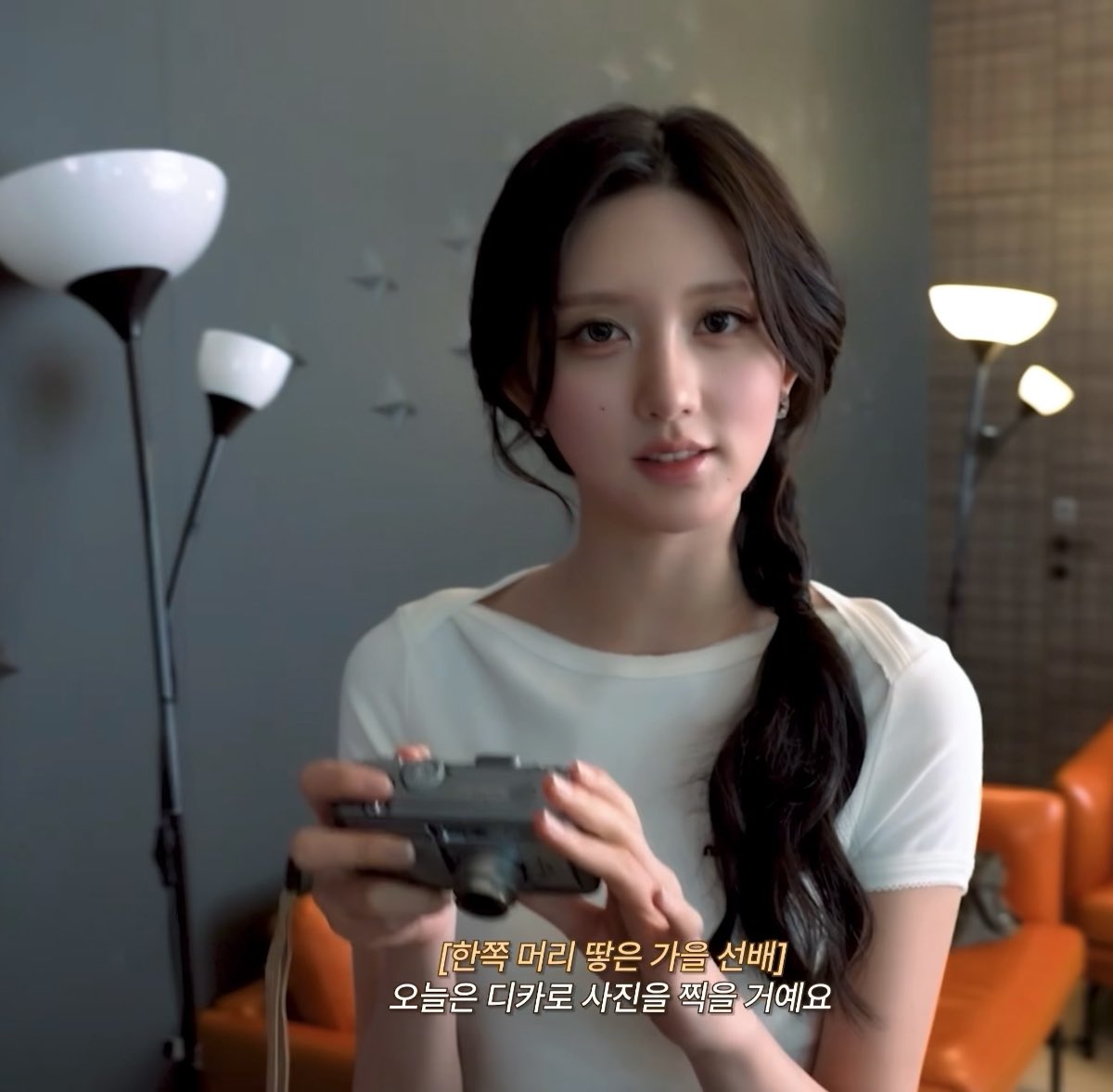 A young woman with long dark hair holding a camera, wearing a white t-shirt, with Korean subtitles reading '[한쪽 머리 땋은 가을 선배] 오늘은 디카로 사진을 찍을 거예요'.