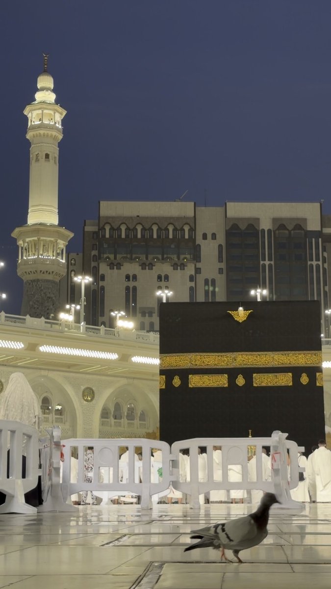 A nighttime view of the Kaaba at Masjid al-Haram in Mecca, with a minaret in the background and pilgrims behind barricades, featuring a date stamp reading '18 Nov 2024'.