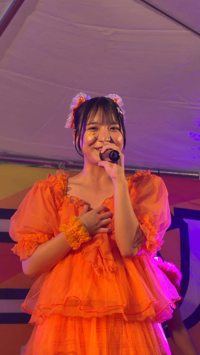 A young woman with a star drawn on her cheek, wearing an orange ruffled dress and hair accessories, singing into a microphone on a stage with pink and purple lighting.