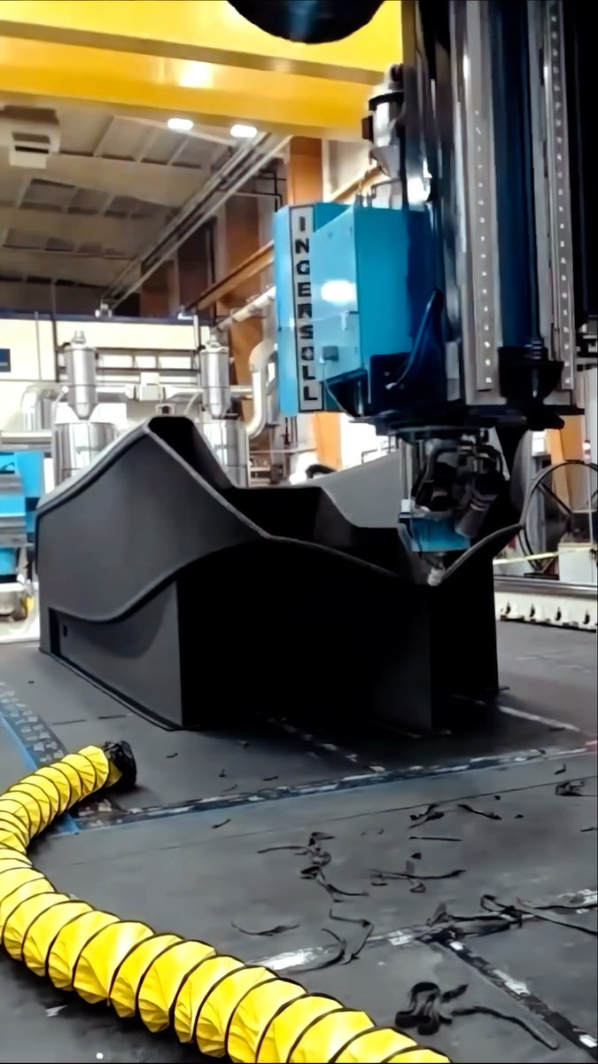 An industrial manufacturing machine cuts a large black plastic component, with black shavings scattered on the floor next to a yellow ventilation hose.