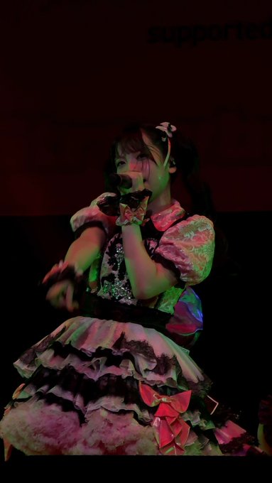 A performer in a frilly pink and black dress with twin pigtails sings into a microphone on a dimly lit stage, with the word "supported" partially visible in the background.