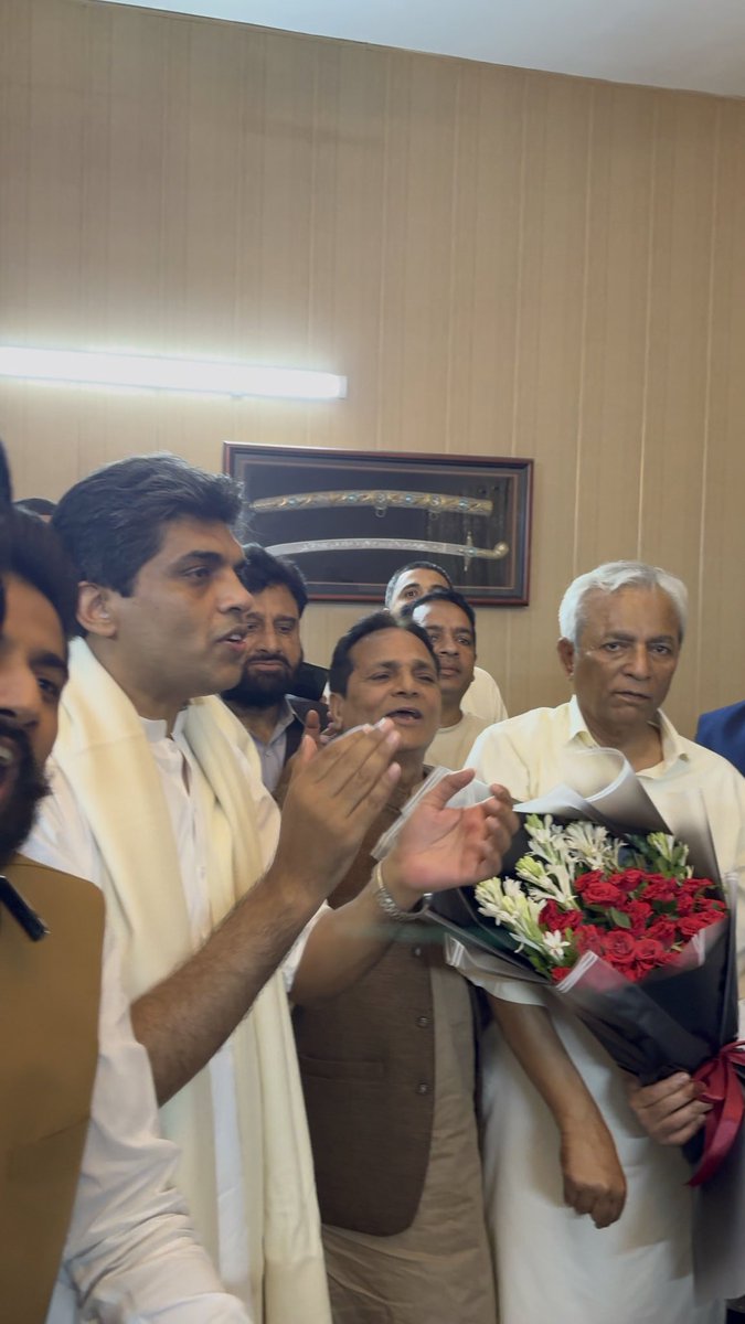 A group of men are gathered indoors, with one clapping and another holding a large bouquet of red roses wrapped in black paper, in front of a wall displaying a framed sword.