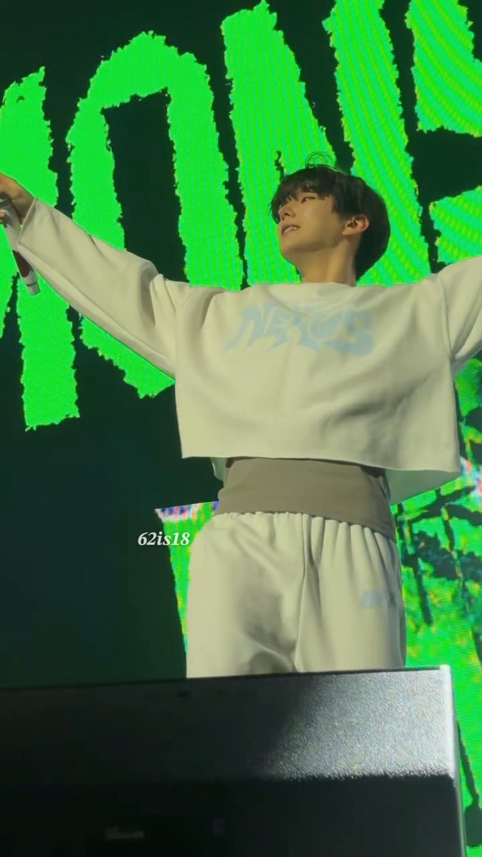 A young male performer on stage wearing a white cropped sweatshirt with light blue text, arms outstretched against a bright green screen backdrop, with a watermark reading '62is18'.