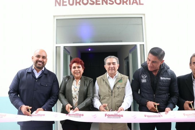 A group of people stand together holding large scissors and a ribbon for a ribbon-cutting ceremony in front of a building labeled 'NEUROSENSORIAL' and 'Familias'.