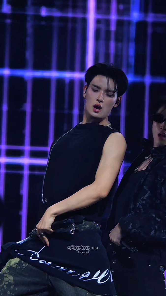 A male performer on stage wearing a black sleeveless top and camouflage pants with 'Courreges' text, performing under purple grid lighting with a watermark '90sKatz'.