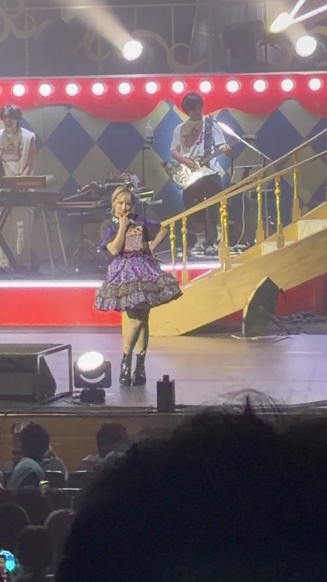 A live concert performance featuring a blonde female vocalist in a frilly purple dress on stage, with a guitarist and keyboardist visible in the background under stage lights.