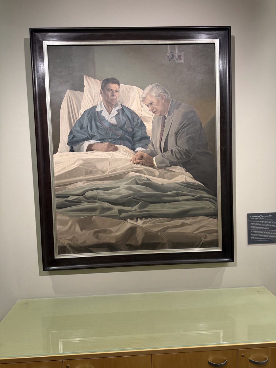 A framed painting displayed in a gallery shows a man lying in a hospital bed while an older man in a suit sits beside him. A small wall label with text is mounted beside the painting, above a glass-topped wooden cabinet.