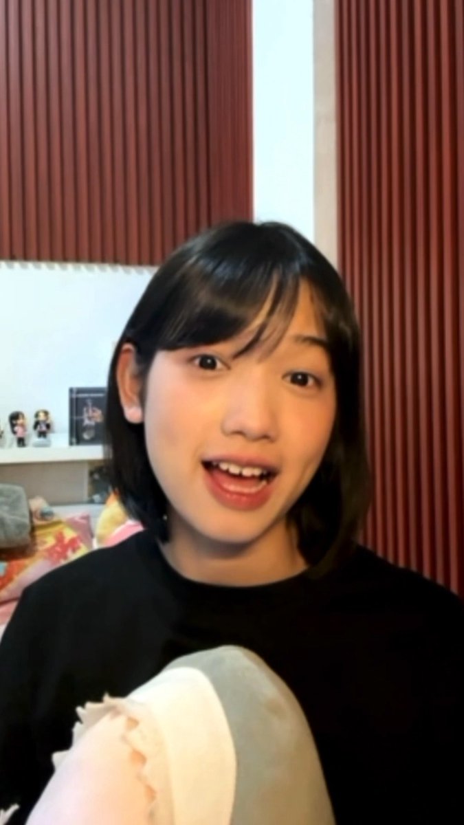 A young woman with shoulder-length dark hair and bangs smiles at the camera while holding a plush toy, standing in a room with red vertical blinds and a shelf of figurines behind her.