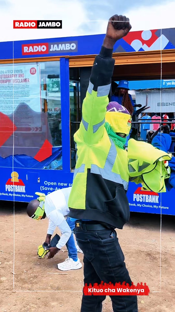 A photo of a person in a high-visibility yellow and black jacket and a colorful face covering raising their fist in front of a blue 'Radio Jambo' and 'PostBank' kiosk, with the caption 'Kituo cha Wakenya' at the bottom and 'Radio Jambo' branding at the top.