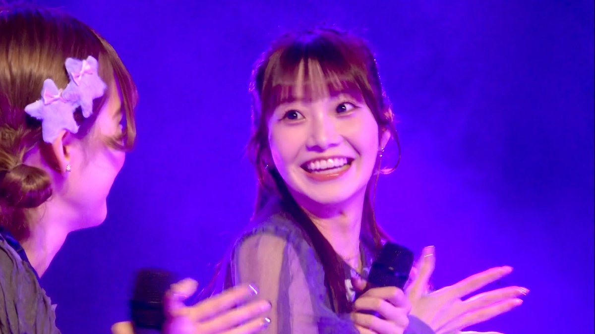 A performance or fan event photo of two smiling young women holding hands against a purple-lit background; one has her hair braided with white bows, and the other is holding a microphone.
