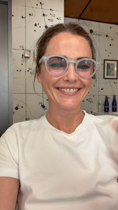 A woman with light blue glasses smiles brightly in a selfie, with a decorative wall behind her displaying numbered celestial-style constellations. The "Books" shelf label visible in the background reinforces the literary and show-related theme of the Felicity Netflix announcement.