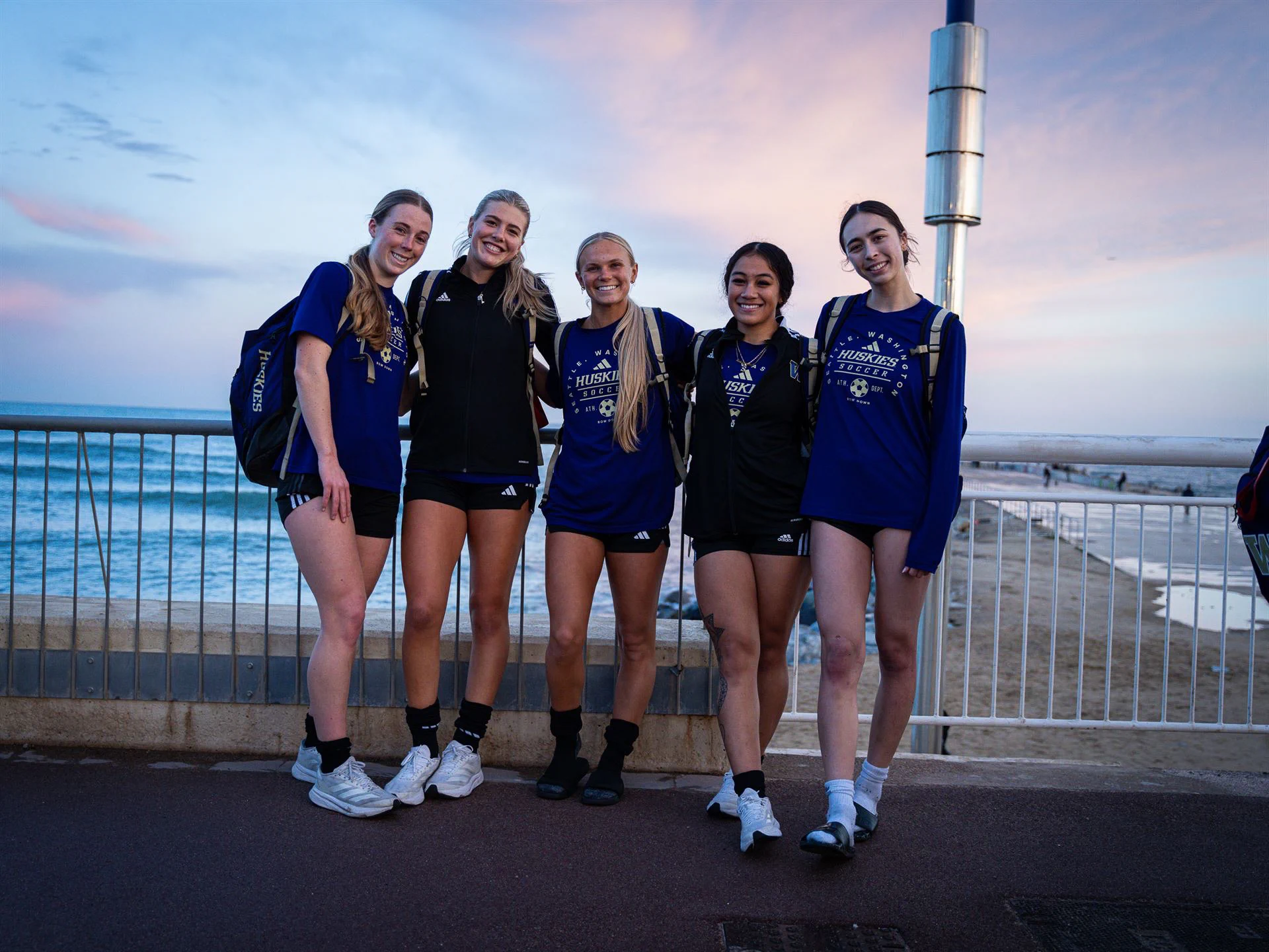 Five smiling young women, likely soccer players, stand shoulder-to-shoulder on a promenade overlooking the ocean at dusk. They wear blue 'HUSKIES SOCCER' shirts, black shorts, and athletic shoes. A metal railing is behind them.