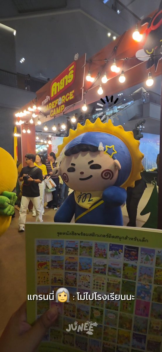 A photo taken at an event featuring a large blue mascot with a sunflower headpiece, with 'ค่ายการ์ตูน' and 'CAREFORCE CAMP' signs visible above.