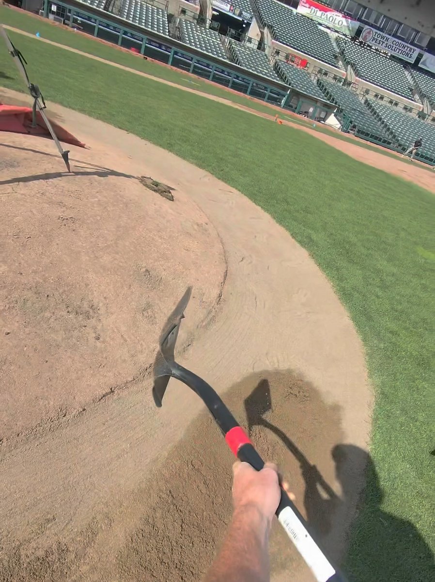 First-person perspective of a groundskeeper holding a pickaxe while working on the dirt infield of a baseball stadium, with empty green seats and advertising banners visible in the background.