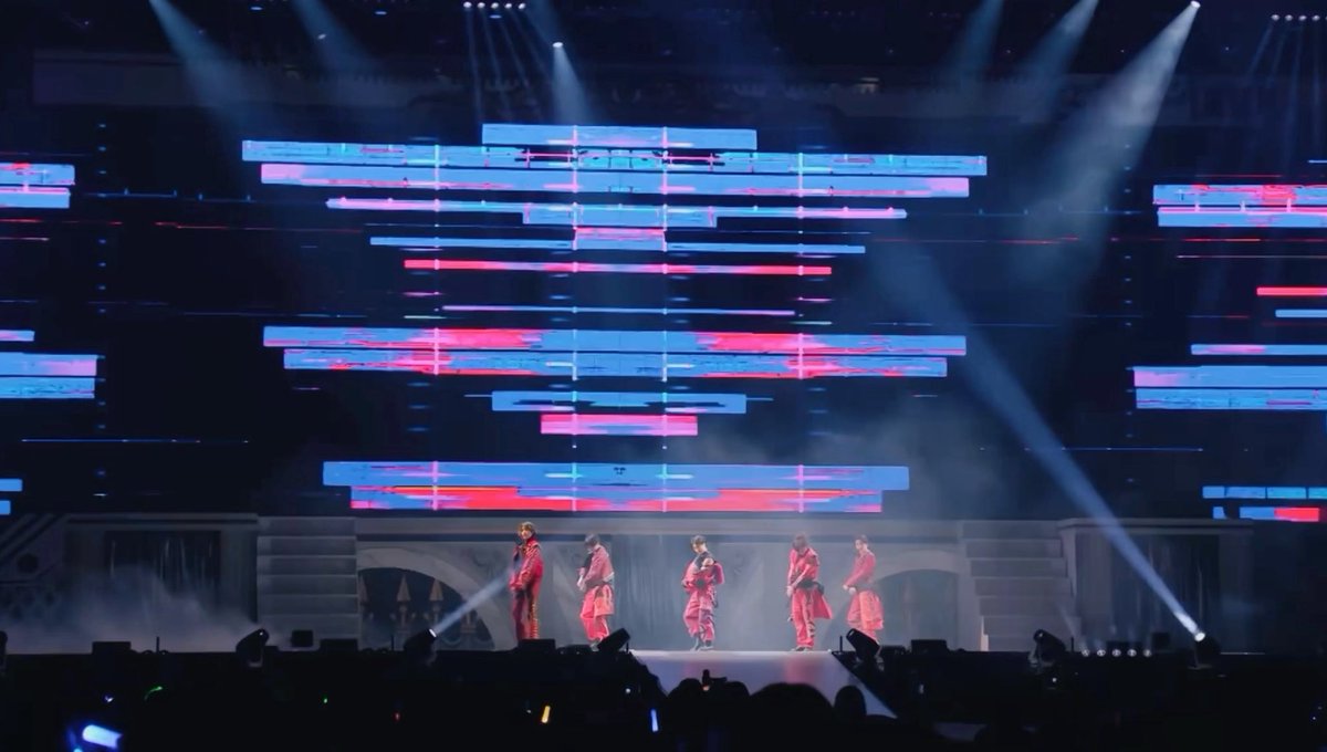A concert performance featuring five performers in red outfits on stage with dramatic lighting and large LED screens displaying abstract blue and red visuals.
