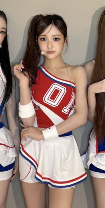 A young woman with brown pigtails wearing a red, white, and blue cheerleader outfit with the number '0' on the front, posing with her arms crossed, with other cheerleaders partially visible beside her.
