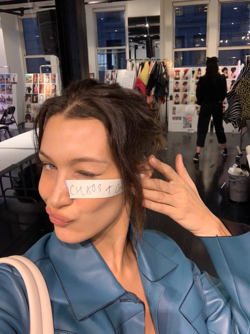 A woman takes a selfie with her eyes closed and a paper taped across her nose reading 'CHAOS +', set in what looks like a fashion studio with reference photos in the background.