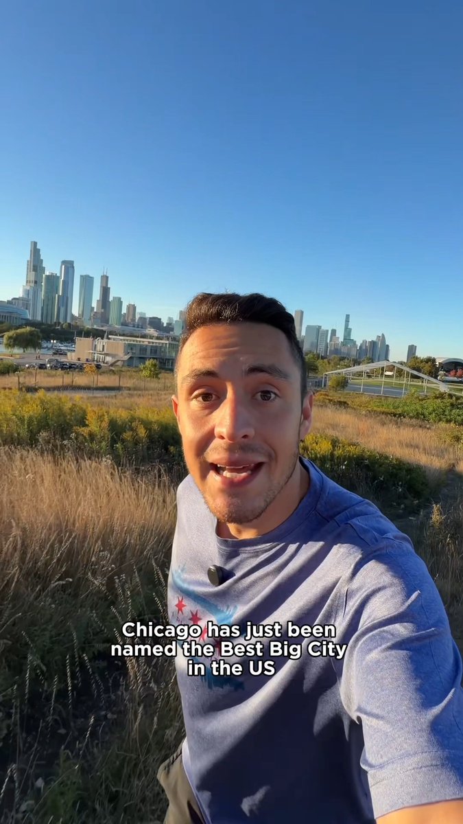 A selfie video of a man in a Chicago-themed shirt speaking to camera with the Chicago skyline in the background, with text overlay reading 'Chicago has just been named the Best Big City in the US'.
