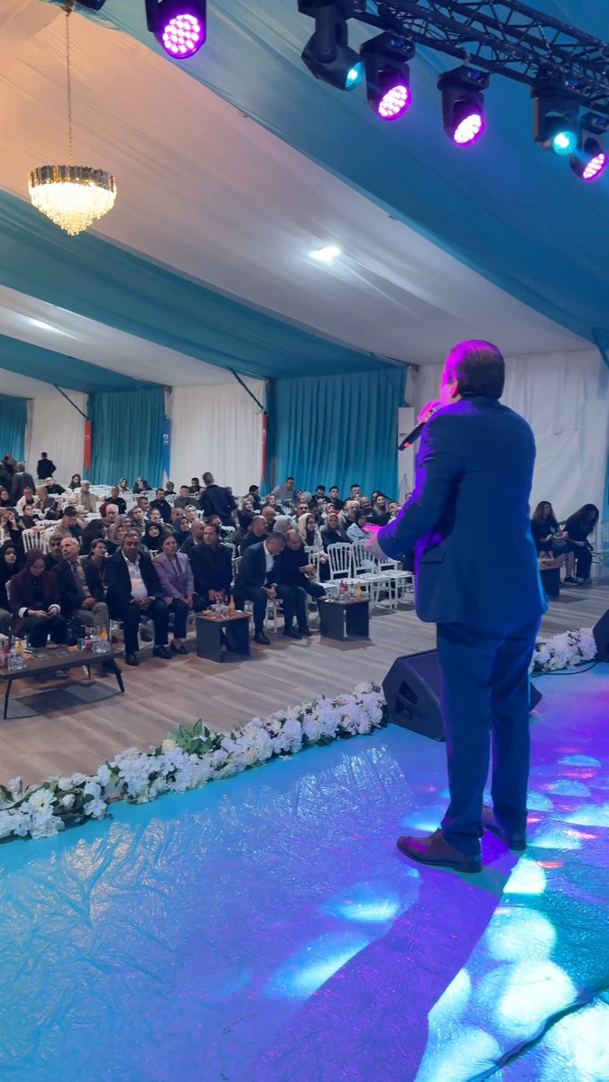 A man in a blue suit stands on a stage with colorful lighting addressing a seated audience at what appears to be an indoor event under a tent with chandeliers.