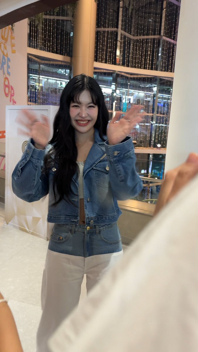 A young woman with long dark hair wearing a denim jacket and jeans smiles and waves at the camera in what appears to be a mall or shopping center.