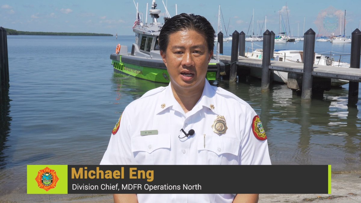 Michael Eng, identified by an on-screen graphic as 'Division Chief, MDFR Operations North,' stands in a white uniform with badges, speaking in front of a green and white fire rescue boat docked at a marina.