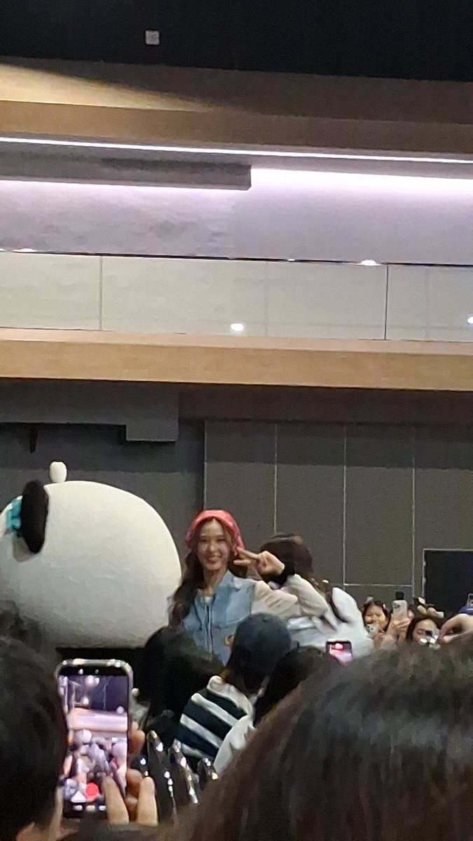 A concert or fan meeting photo showing a woman with a pink headband posing with a peace sign, accompanied by someone in a panda costume, while fans in the foreground hold up phones to take pictures.