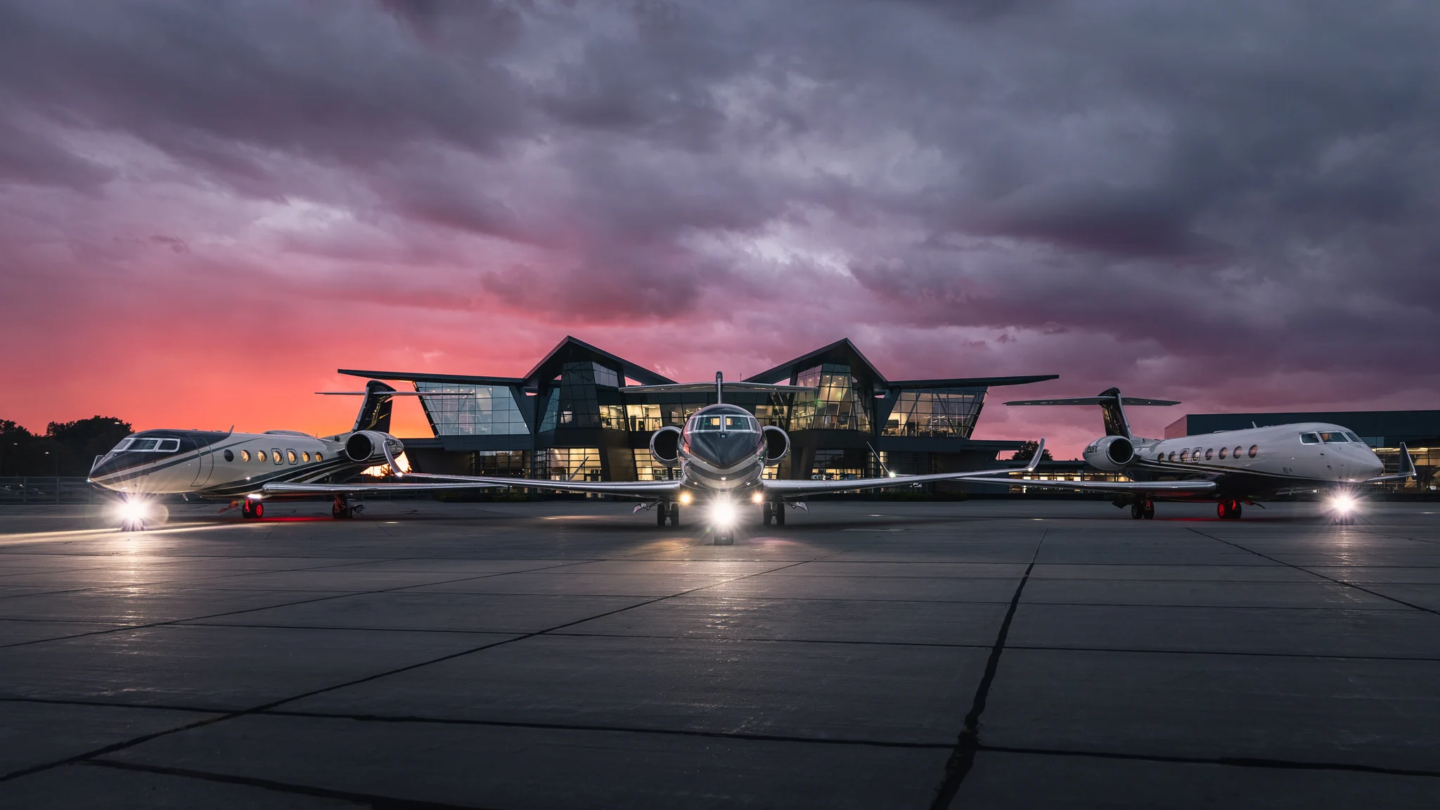 Three sleek private jets are parked on an airport tarmac at dusk, with a modern terminal building in the background. The sky is a blend of purple and pink hues, creating a dramatic sunset scene.