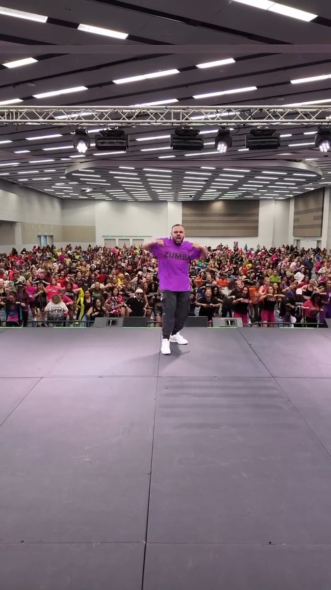 An energetic Zumba instructor in a purple shirt dancing on a large stage in front of a massive crowd of participants inside a spacious convention hall or arena.