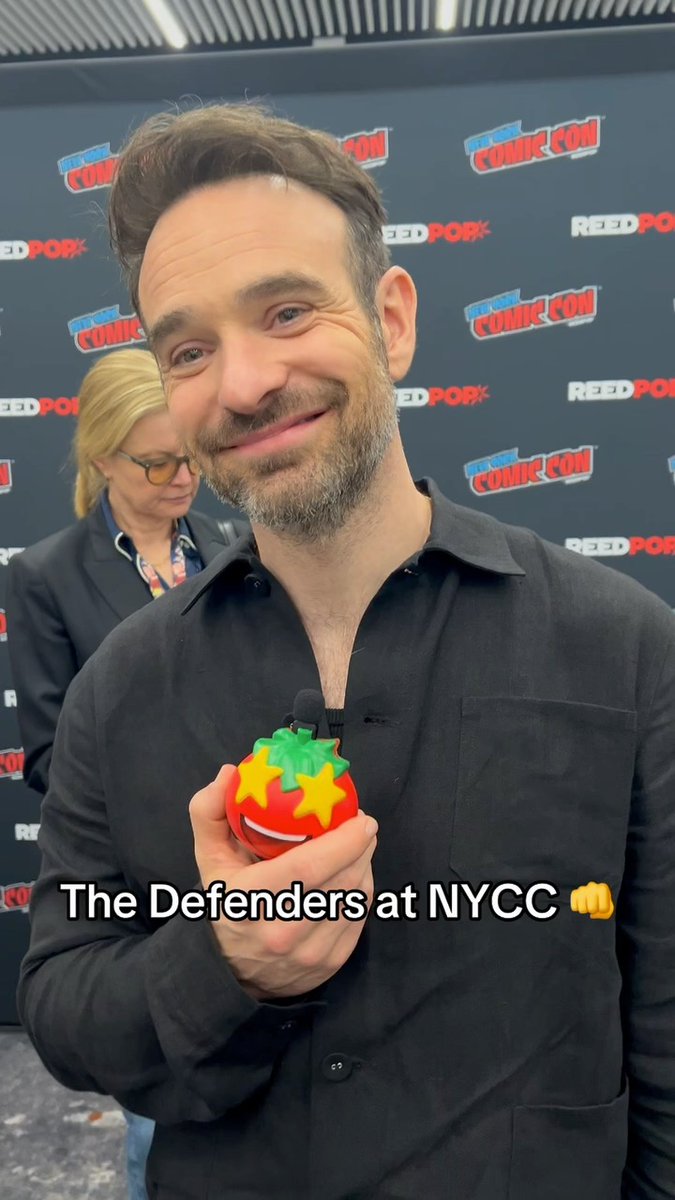 A man in a black button-up shirt smiles while holding a red plush tomato with star eyes at New York Comic Con, with "The Defenders at NYCC" overlaid on the image and Comic Con/ReedPop logos visible on the backdrop behind him.