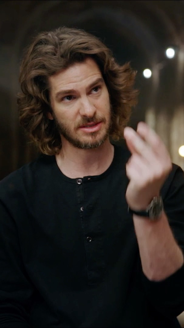 A man with curly brown hair and a beard, wearing a black henley shirt, gestures with his right hand while wearing a watch.