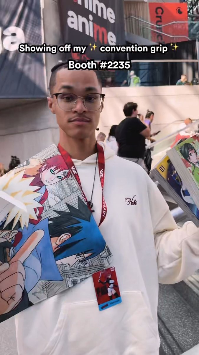 A young man wearing glasses and a cream hoodie proudly displays manga volumes at an anime convention booth, with a lanyard and badge hanging around his neck. The image includes a text overlay reading "Showing off my ✨ convention grip ✨ Booth #2235," and a Crunchyroll "anime" banner is visible in the background, tying directly to the post's promotion of booth shopping and exclusive merchandise.