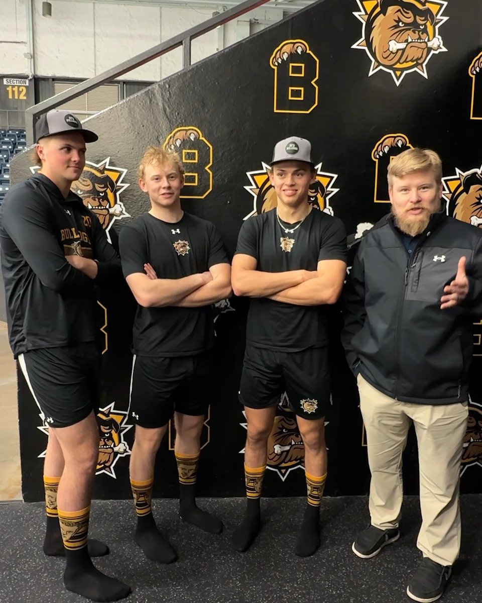 Three young men in matching black 'Bulldogs' athletic wear posing with a bearded man in a gray jacket at what appears to be a sports venue with a Bulldogs logo backdrop.