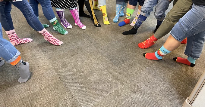 A group of people standing in a circle, each wearing colorful and uniquely patterned socks, including stripes, polka dots, and cartoon prints. The floor is carpeted in a neutral tone.