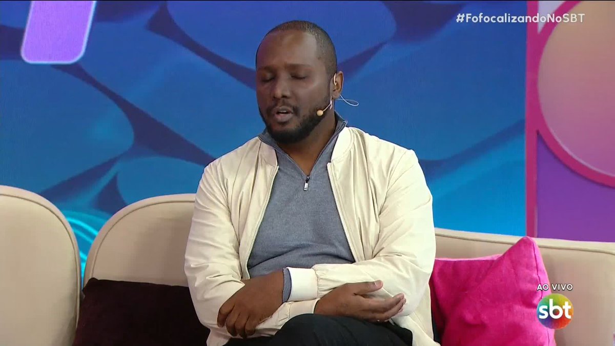 A man in a white jacket and gray shirt sits on a couch with his eyes closed on the SBT show 'Fofocalizando' (hashtag #FofocalizandoNoSBT), broadcast 'ao vivo' (live).