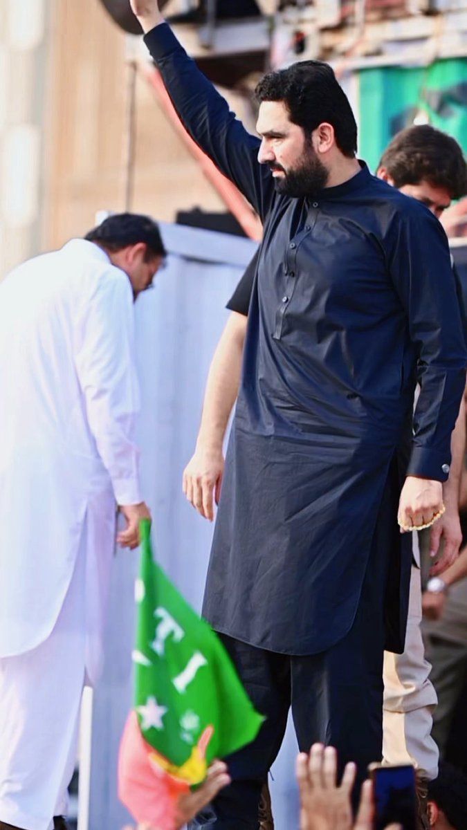 A man with a beard wearing a black kurta raises his arm in front of a crowd, with someone holding a green flag visible at the bottom of the frame.