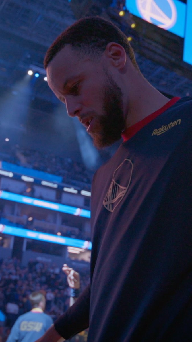 Stephen Curry, wearing a Golden State Warriors warmup shirt with the Rakuten logo, looks down thoughtfully while standing in a brightly lit arena, likely captured during a documentary-style moment from the IMAX film "Portrait of an Artist: Stephen Curry." The image showcases the iconic player reflecting before a crowd, tying directly to the promotional post celebrating the release of his biographical film.