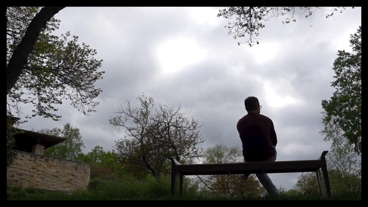 A silhouette of a person wearing glasses and a maroon sweater sitting on a wooden bench, looking up at a cloudy sky framed by trees.
