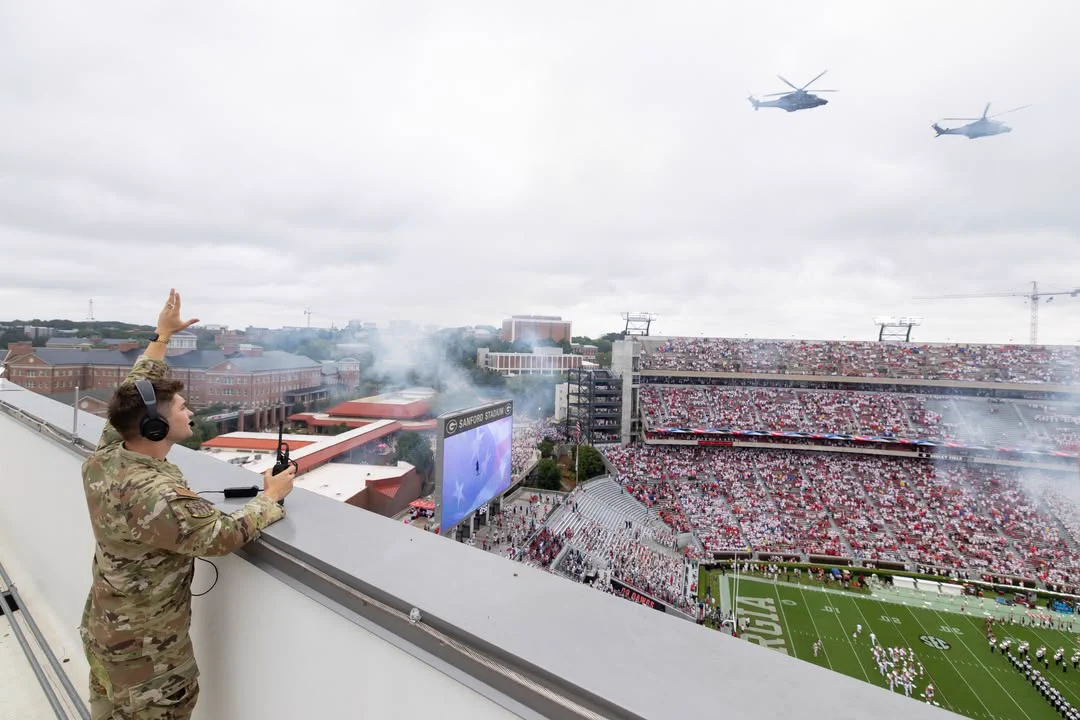 A person in military uniform, standing on a rooftop overlooking a large sports stadium, gestures upward as two helicopters fly by. The stadium is filled with spectators and features a sports field.