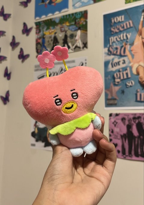 A close-up photo of a hand holding a small, pink plush toy with a yellow beak and two pink flower antennae, set against a wall decorated with posters and butterfly cutouts.