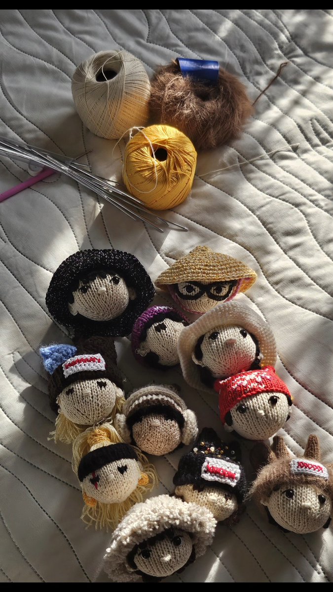 A collection of small knitted dolls with various hats and accessories, along with balls of yellow and beige yarn and knitting needles, arranged on a white quilted surface.