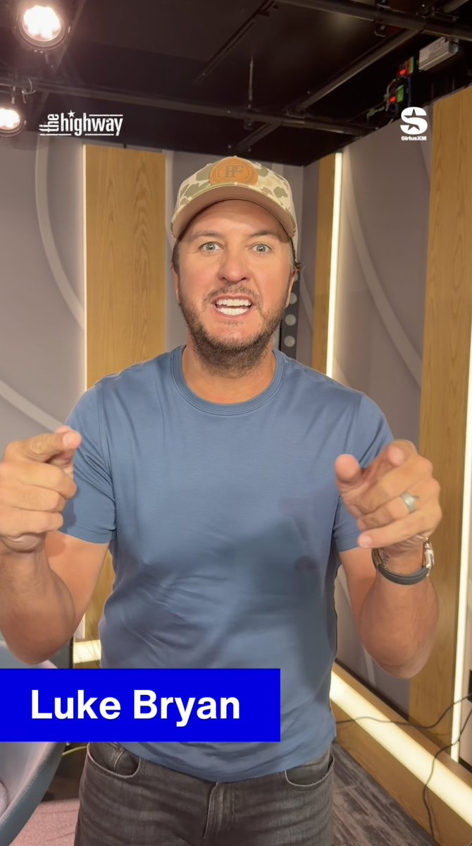 Country music star Luke Bryan, identified by a text overlay, stands in a studio wearing a blue t-shirt and a camouflage hat with an orange 'H' logo, pointing towards the camera while 'THE HIGHWAY' and SiriusXM logos appear at the top.