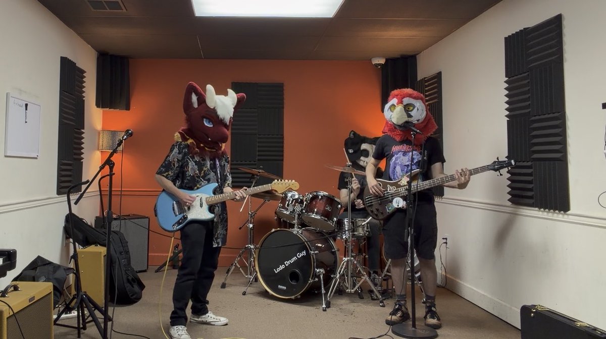 Three people wearing mascot heads (a red dragon and a red eagle/bird) playing electric guitars and drums in a soundproofed practice room with acoustic foam panels.