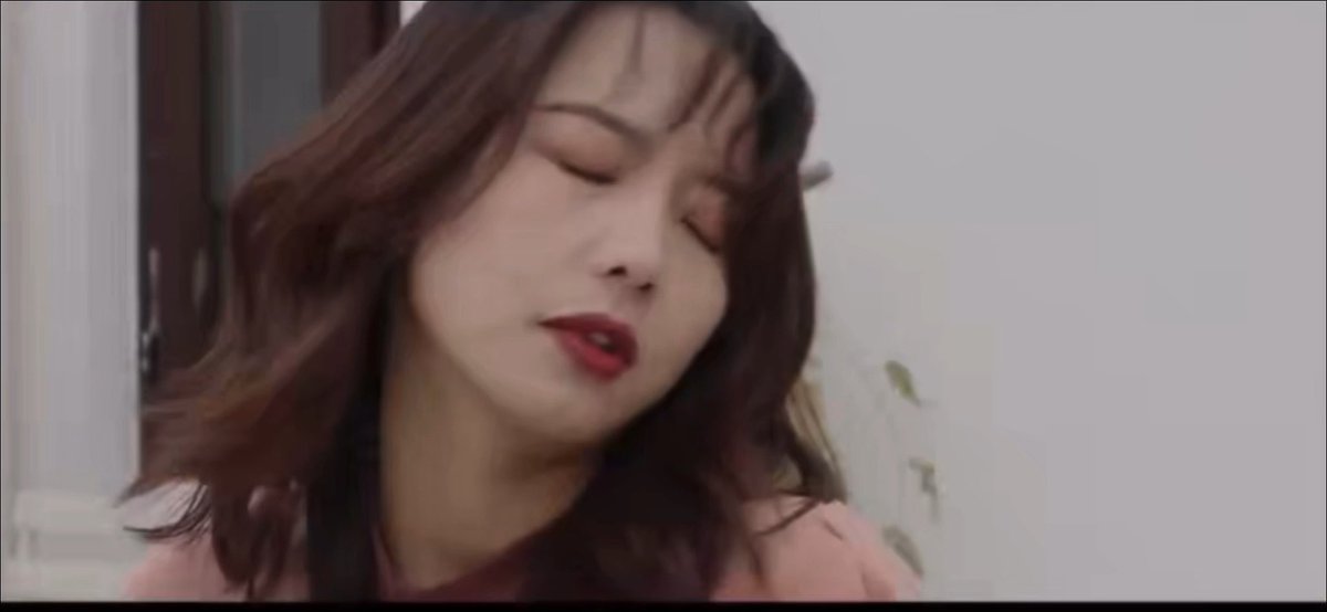 A screenshot from a video showing a woman with long dark wavy hair and red lipstick, her eyes closed and lips slightly parted.