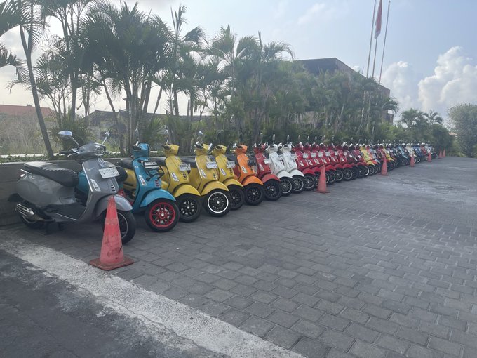 A row of colorful scooters lined up in a parking lot in front of a building with palm trees.