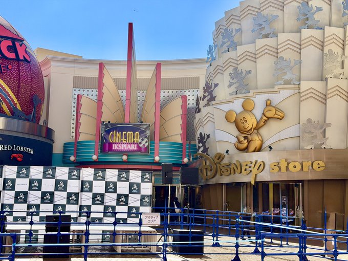 An exterior view of an Ikspiari entertainment complex featuring a 'Cinema IKSPIARI' sign, a 'Disney Store', and a large Mickey Mouse figure near the entrance.