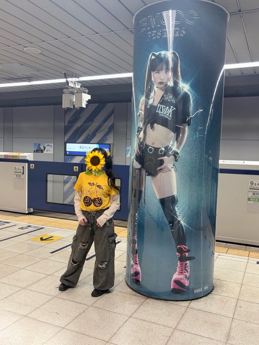 A person in a yellow sunflower-print shirt and baggy jeans poses in a subway station next to a large pillar advertisement featuring a performer in black leather.