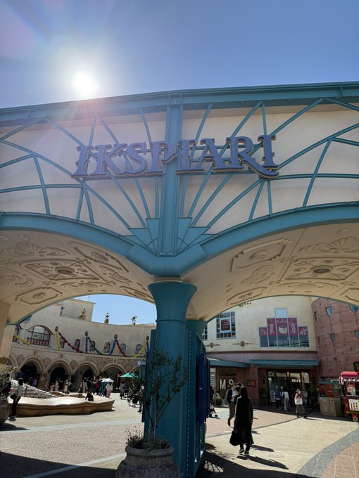An outdoor entrance to 'IKSPIARI,' a shopping and entertainment complex, featuring a large blue and white archway with the name displayed prominently under a bright sun.
