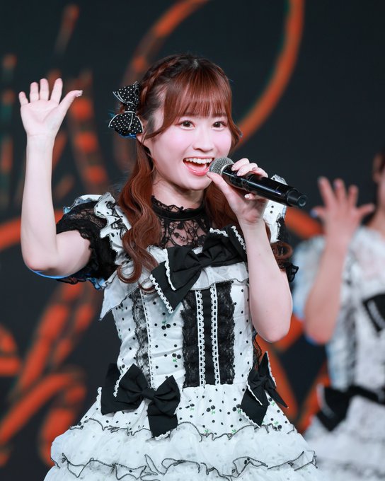 A young woman with reddish-brown hair styled with a black bow smiles and waves while singing into a microphone, wearing a white and black frilly performance dress on stage.